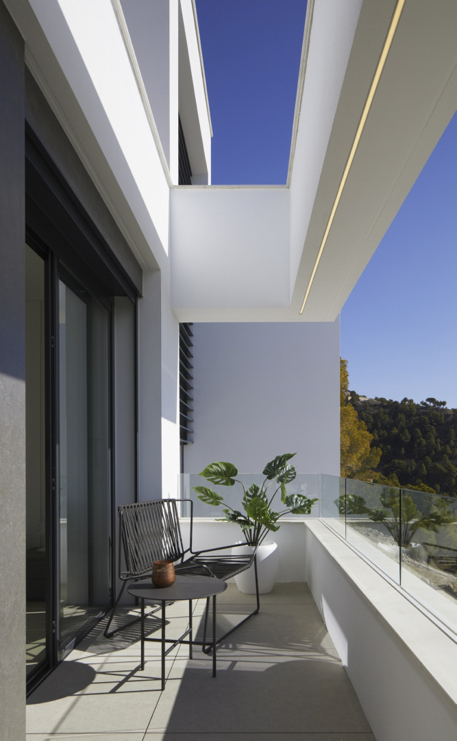 Private Terrasse mit Glasgeländer, minimalistischen Außenmöbeln und Bergblick. Der weiße Dachüberstand verfügt über eine integrierte lineare LED-Beleuchtung.