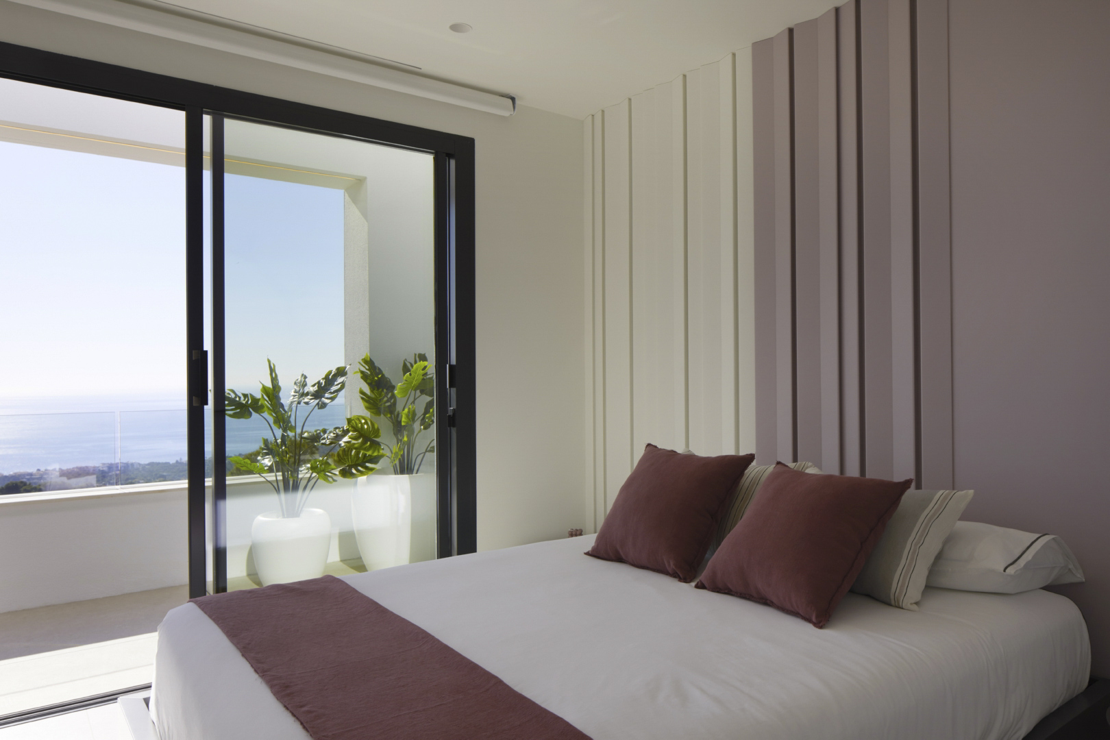 Schlafzimmer mit vertikaler Wandtäfelung und Glasschiebetüren zur Terrasse mit Pflanzkübeln und weitem Blick auf das Mittelmeer.