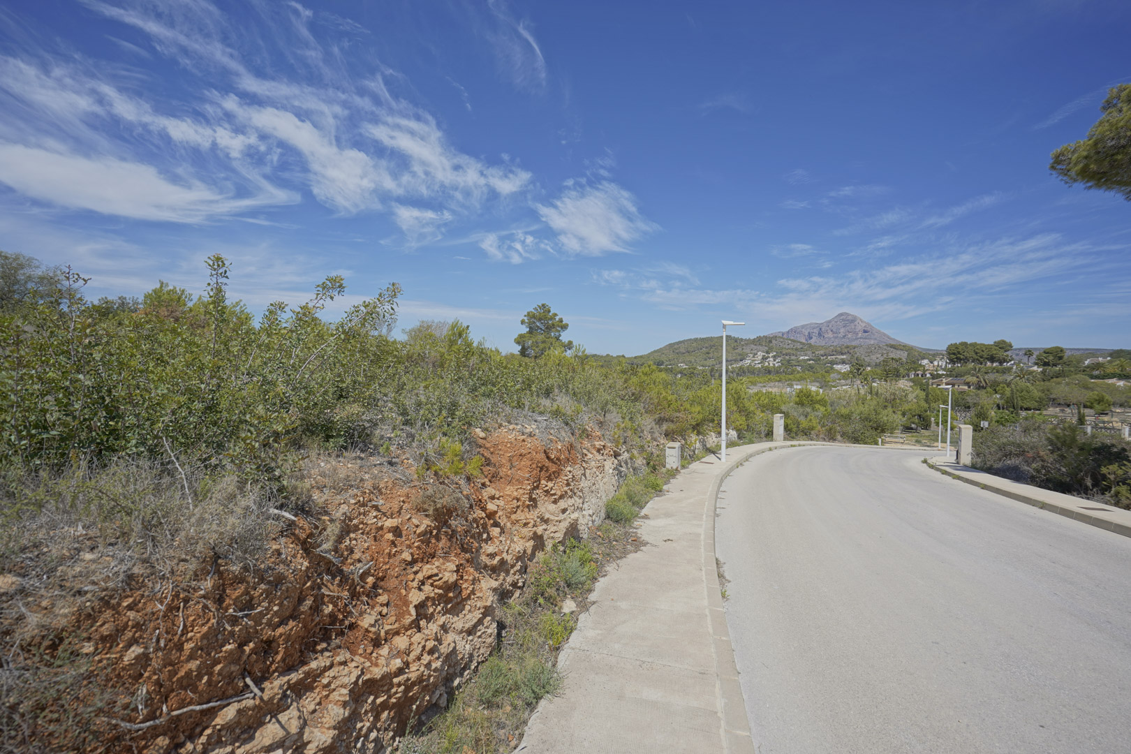 Blick auf die Zufahrtsstraße im Wohngebiet Valsol in Jávea mit dem Berg Montgó im Hintergrund und typisch mediterraner Vegetation.