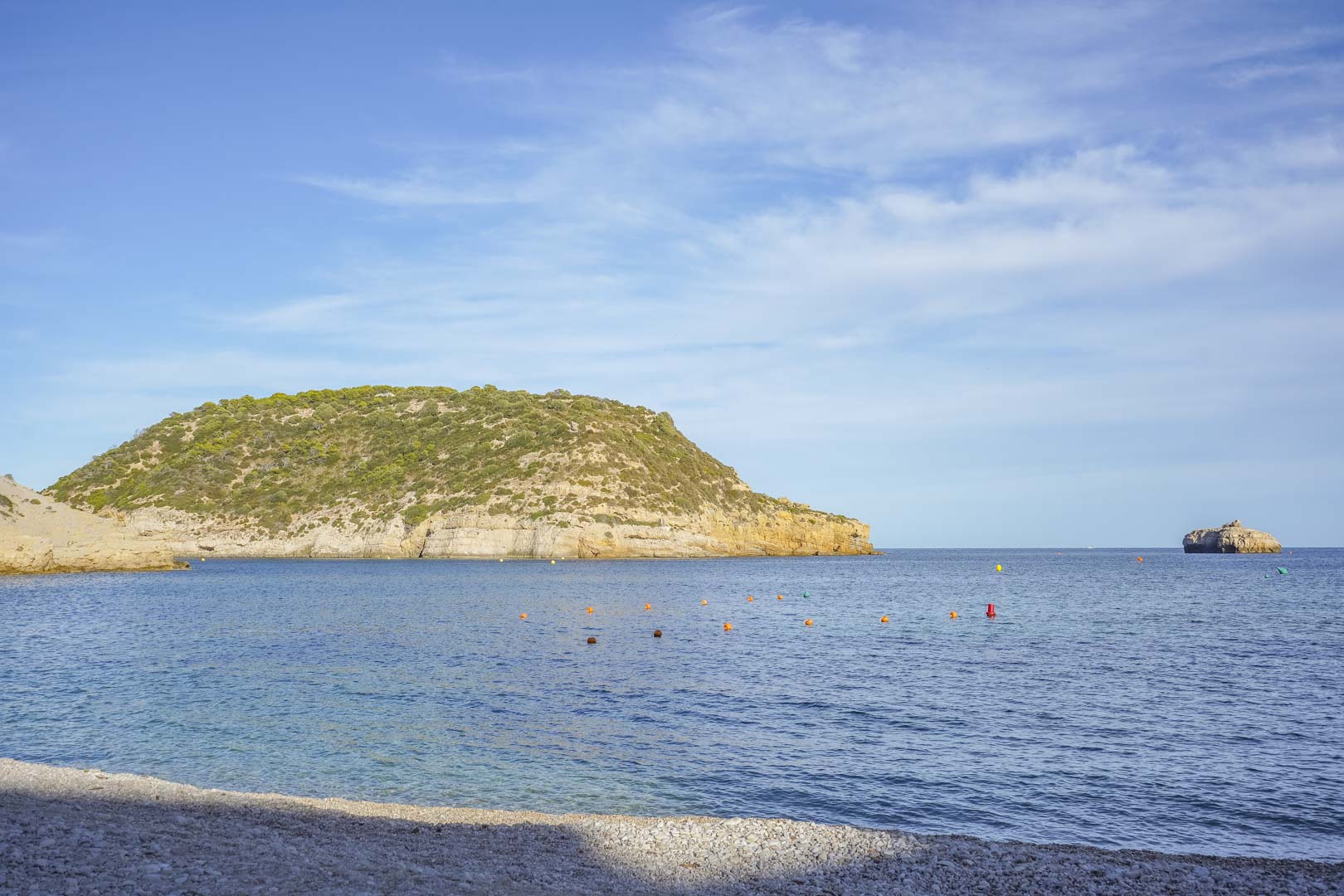 Panoramablick auf einen Kieselstrand in Jávea mit kristallklarem Mittelmeerwasser und markanten Küstenfelsen unter strahlend blauem Himmel.