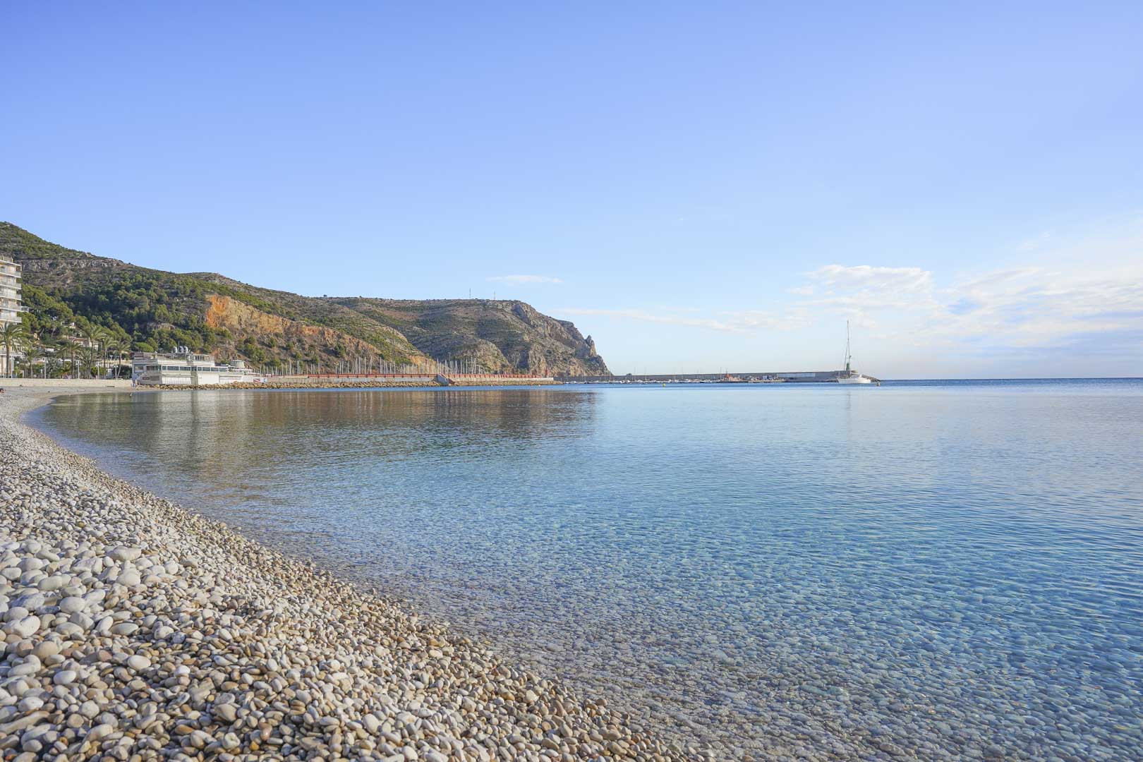 Panoramablick auf einen Kieselstrand in Jávea mit kristallklarem Wasser und den Klippen des Cabo de San Antonio im Hintergrund bei klarem Himmel.