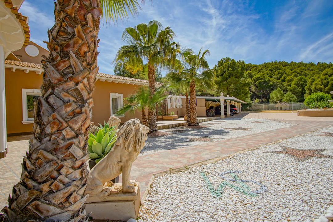 Außenbereich mit Steinlöwen-Statuen, Palmen und einer dekorativen Kieseinfahrt mit Mosaiken, die zu einem überdachten Carport vor einer Waldkulisse führt.