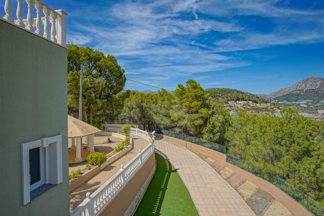 Blick auf die gepflasterte Auffahrt und die klassischen weißen Balustraden. Ein schattiger Pavillon liegt inmitten von Pinien mit weitem Blick auf die Berge.