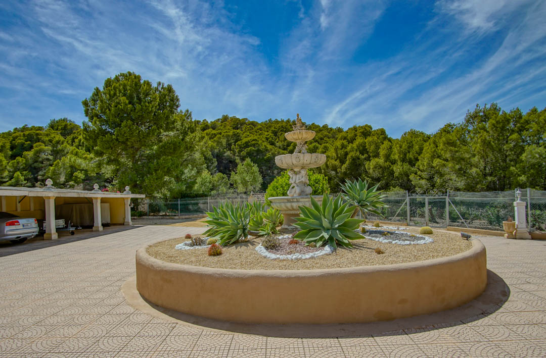 Haupteingang mit dreistufigem klassischem Brunnen und kreisförmigem Beet mit Agaven, angrenzend an einen Carport und Pinienwald.