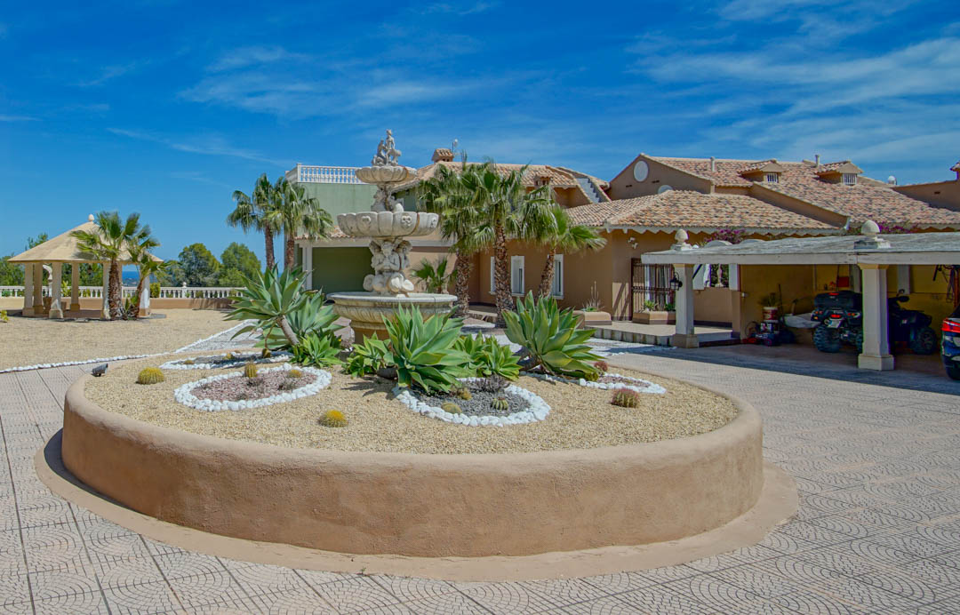 Haupteingang mit ornamentalem Steinbrunnen, rundem Beet mit Agaven und Kakteen, überdachtem Carport und Gartenpavillon vor blauem Himmel.