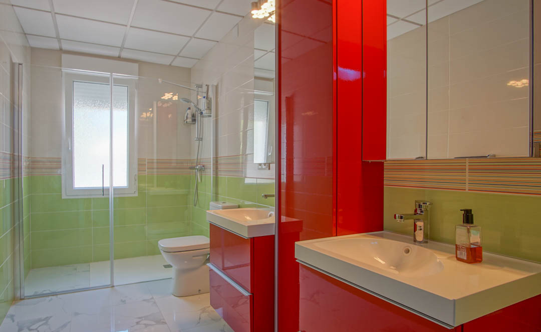 Modernes Badezimmer mit glänzend rotem Waschtisch, begehbarer Glasdusche und hellgrünen Akzentfliesen auf einem Boden in Marmoroptik.