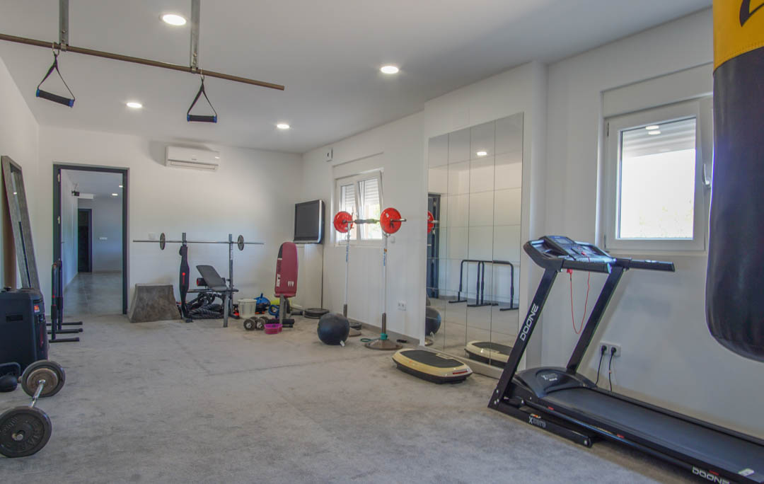 Privater Fitnessraum mit Laufband, Boxsack und Hantelstation. Ausgestattet mit Wandspiegeln, Deckenspots und einer modernen Klimaanlage.