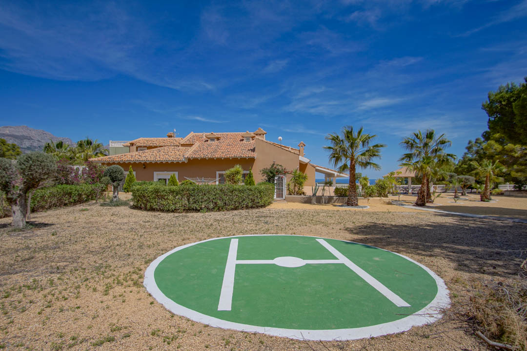 Privater Hubschrauberlandeplatz auf dem weitläufigen Grundstück, umgeben von mediterranen Gärten mit Palmen und Blick auf das Haupthaus mit Terrakottadach.