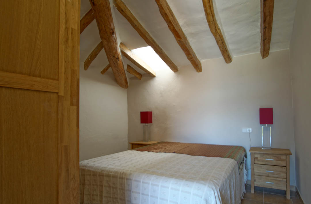 Rustikales Schlafzimmer mit sichtbaren Holzbalken, einem Dachfenster für natürliches Licht und weißen Putzwänden. Mit Nachttischen aus Holz und roten Lampen ausgestattet.