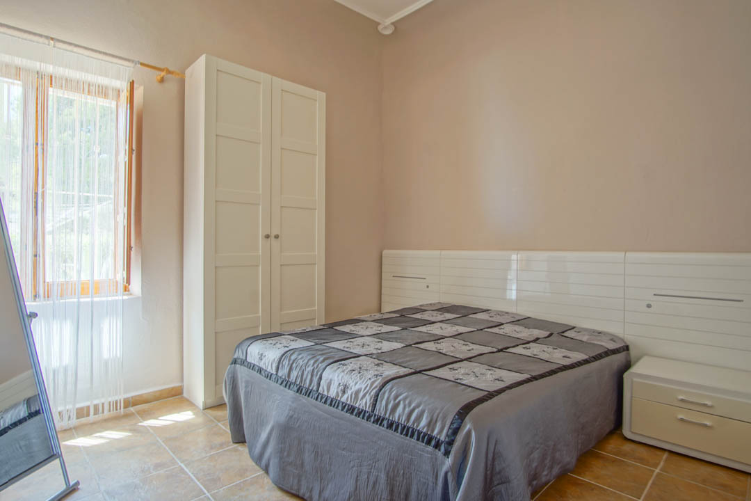 Schlafzimmer mit Keramikfliesenboden, freistehendem weißem Kleiderschrank und modernem Kopfteil mit integrierten Nachttischen und Tageslicht.