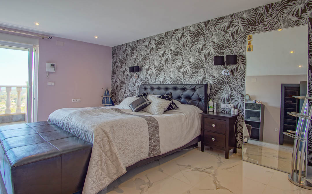 Schlafzimmer mit Marmorboden, Akzentwand in Tropenoptik, gepolstertem Lederkopfteil und direktem Zugang zu einem Balkon mit Balustrade.