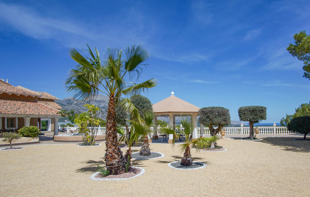 Weitläufiger Kieshof mit Palmen und Formschnittgehölzen, ergänzt durch einen weißen Säulenpavillon mit Panoramablick auf das Meer und die Berge.
