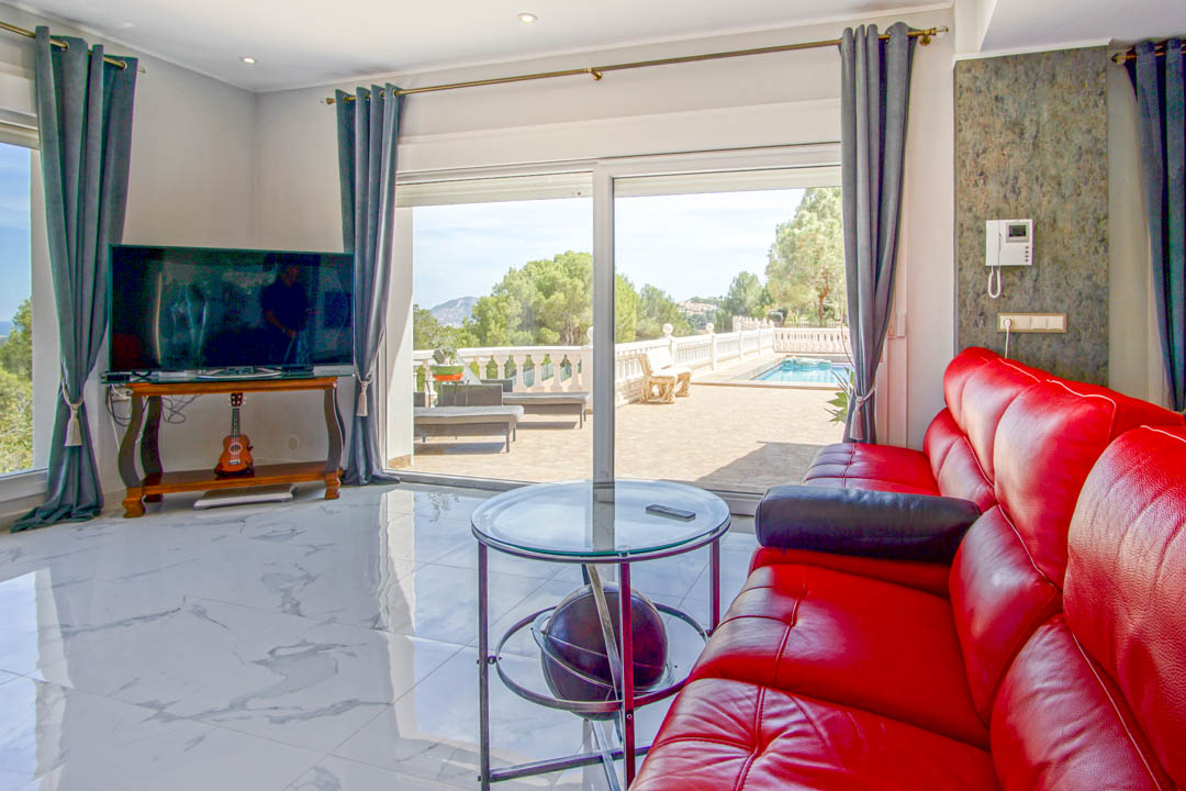 Wohnbereich mit weißem Marmorboden, rotem Ledersofa und großen Glasschiebetüren, die direkt auf die Terrasse mit Pool und Weitblick führen.