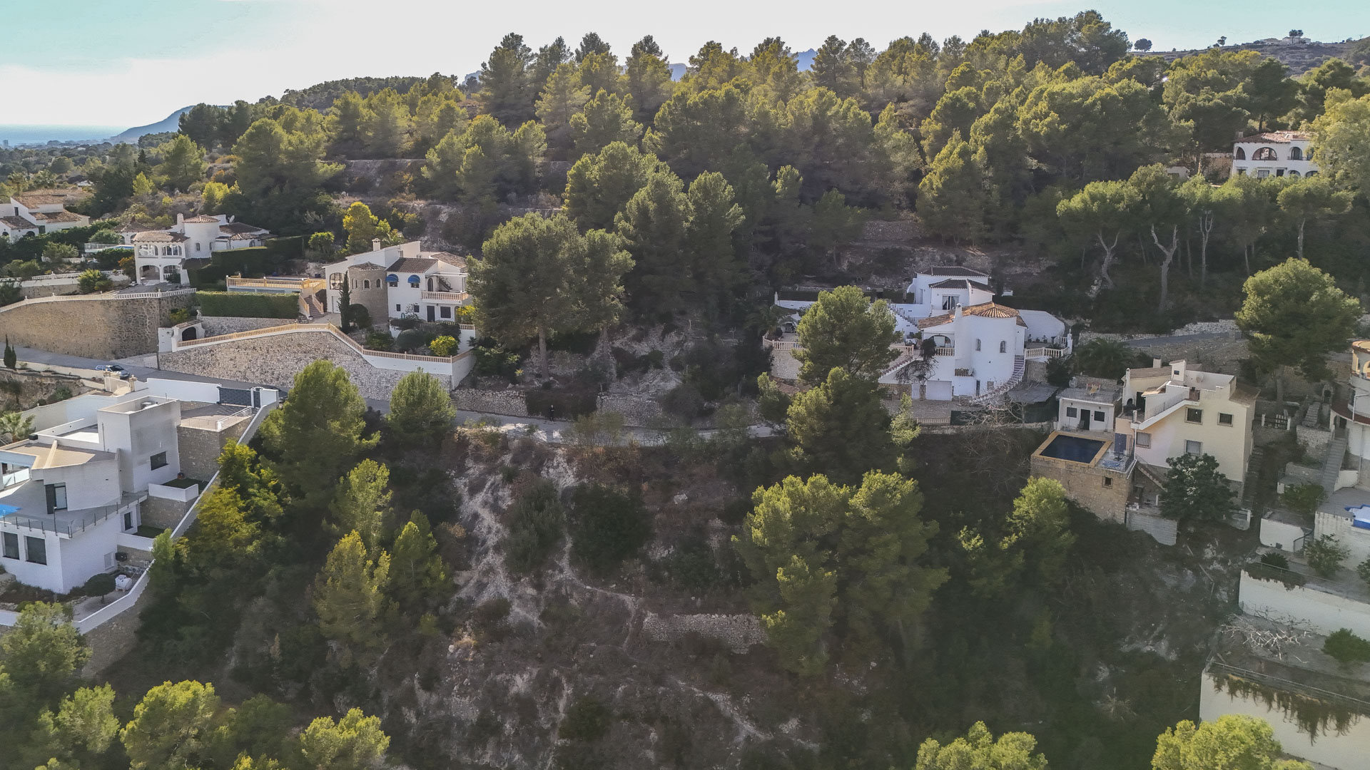 Luftaufnahme von Wohngrundstücken in Benissa, eingebettet in einen dichten Pinienwald mit Natursteinmauern und Blick auf das ferne Mittelmeer.
