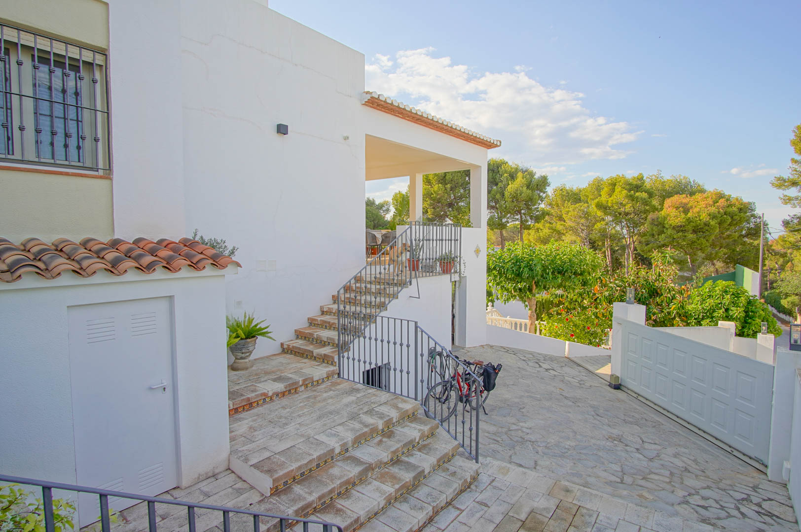 Außenbereich mit gefliesten Treppen und dekorativen Setzstufen, die zur überdachten Terrasse führen, eingebettet in eine mediterrane Gartenlandschaft.