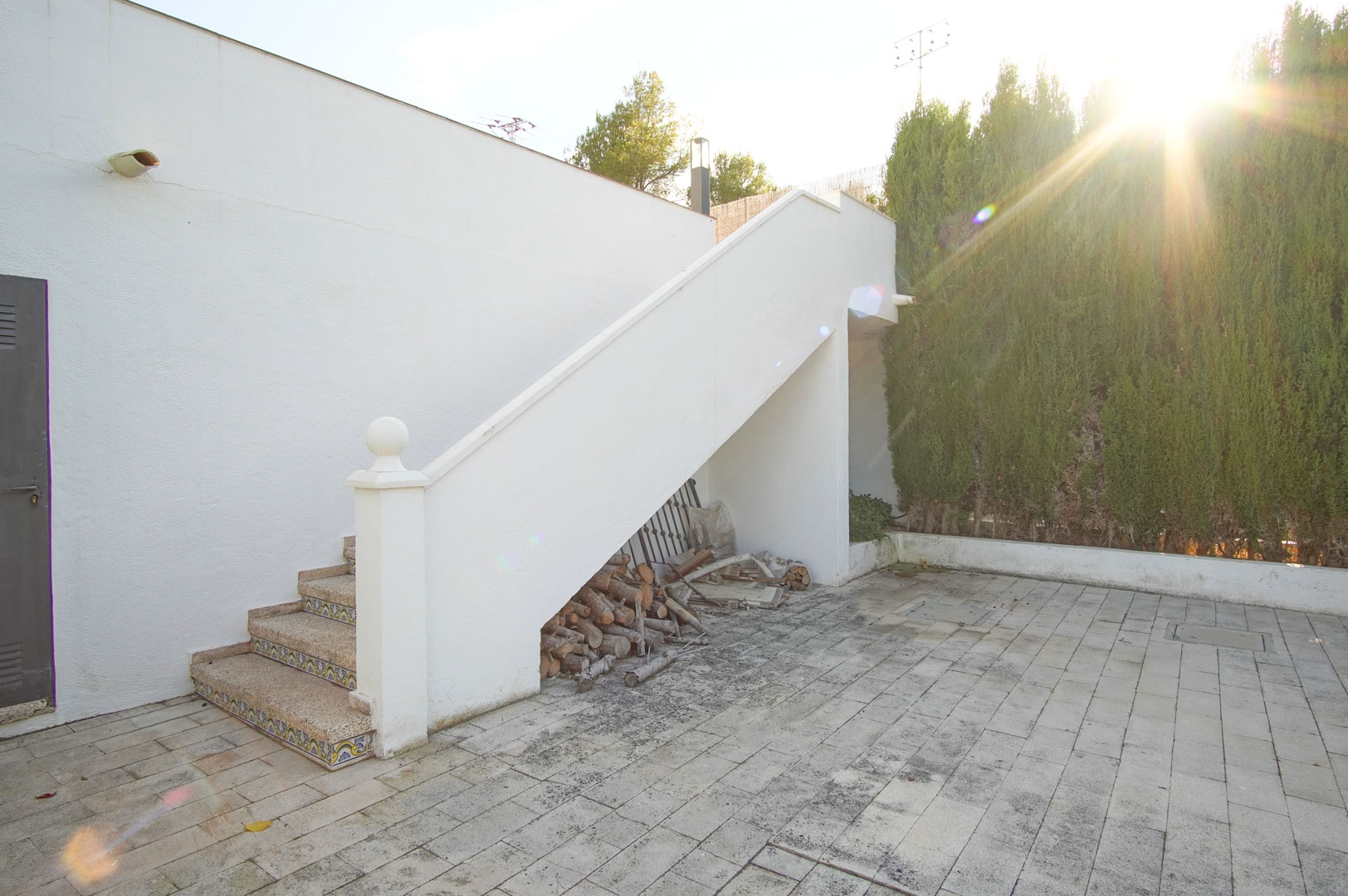 Außenliegende Steintreppe mit dekorativen Fliesendetails, die zu einer oberen Terrasse führt, angrenzend an einen gepflasterten Hof mit Holzlager.