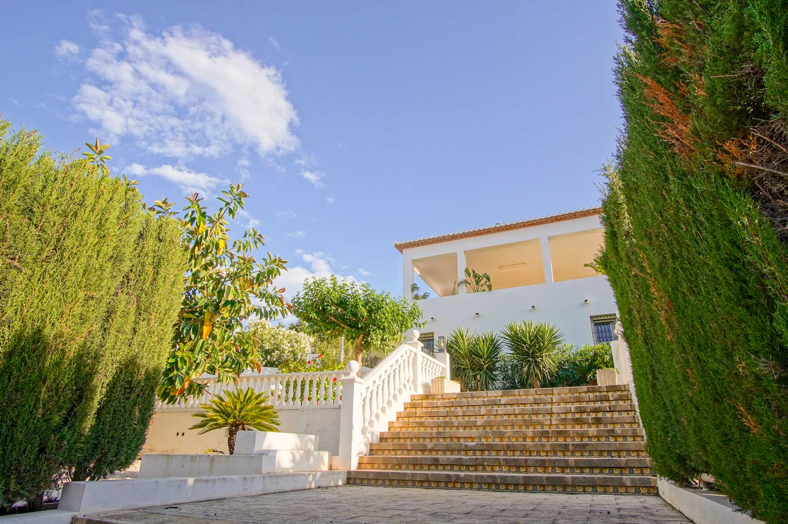 Außentreppe aus Stein mit dekorativen Setzstufen und weißer Balustrade, gesäumt von hohen Zypressenhecken vor der weißen Fassade der Villa.