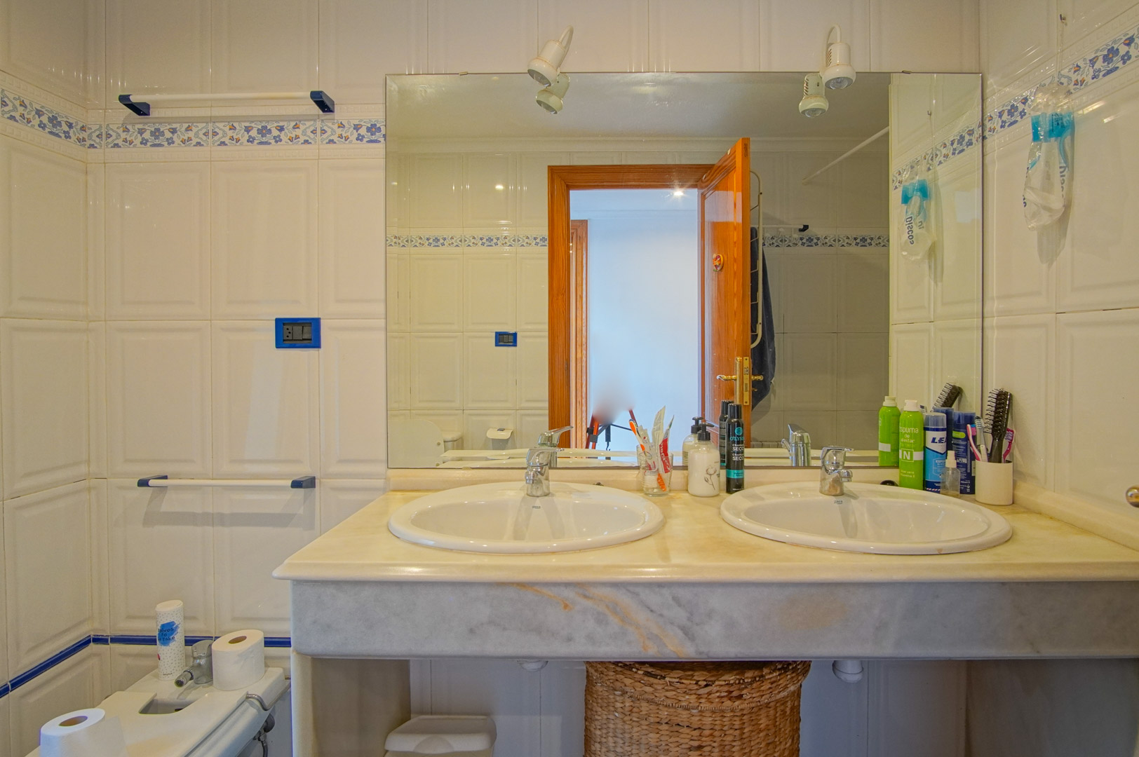 Badezimmer mit Doppelwaschbecken auf Marmorplatte, weißen Fliesen mit blauer Blumendekorleiste und großem Spiegel mit integrierter Beleuchtung.