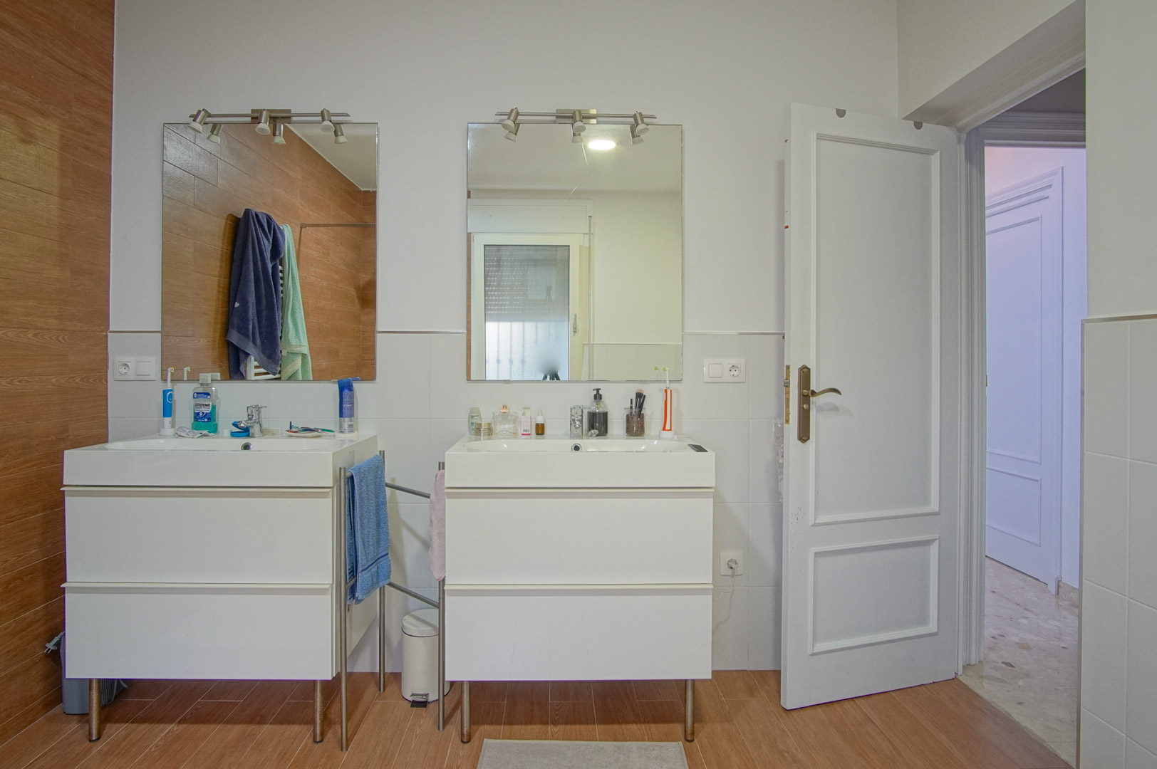 Badezimmer mit Doppelwaschtisch, separaten weißen Unterschränken, beleuchteten Spiegeln und einer Wandgestaltung aus weißen Fliesen und Holzoptik-Elementen.