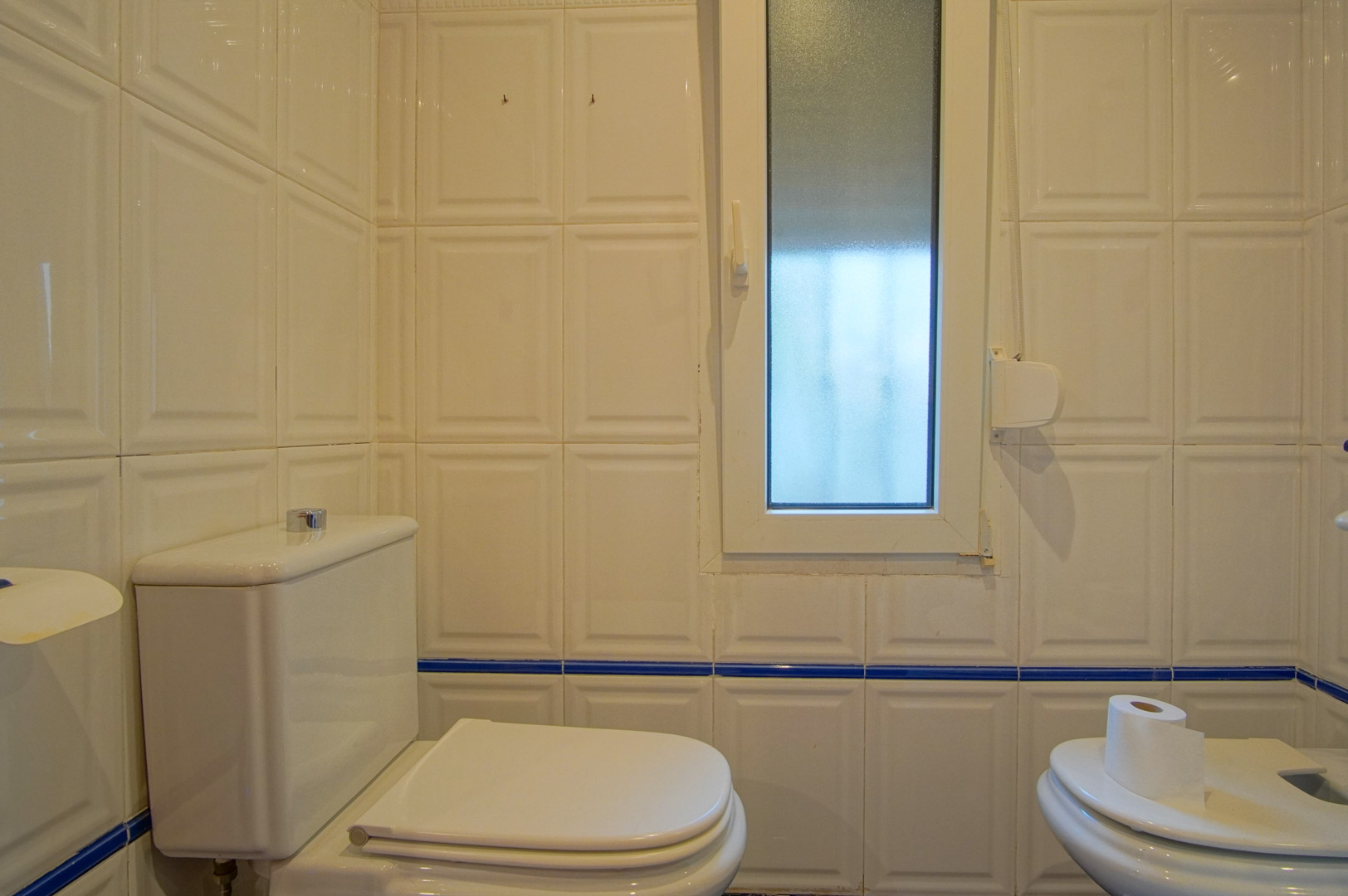 Badezimmer mit weiß reliefierten Wandfliesen und blauer Bordüre. Ausgestattet mit WC, Bidet und einem Milchglasfenster für natürliches Licht.