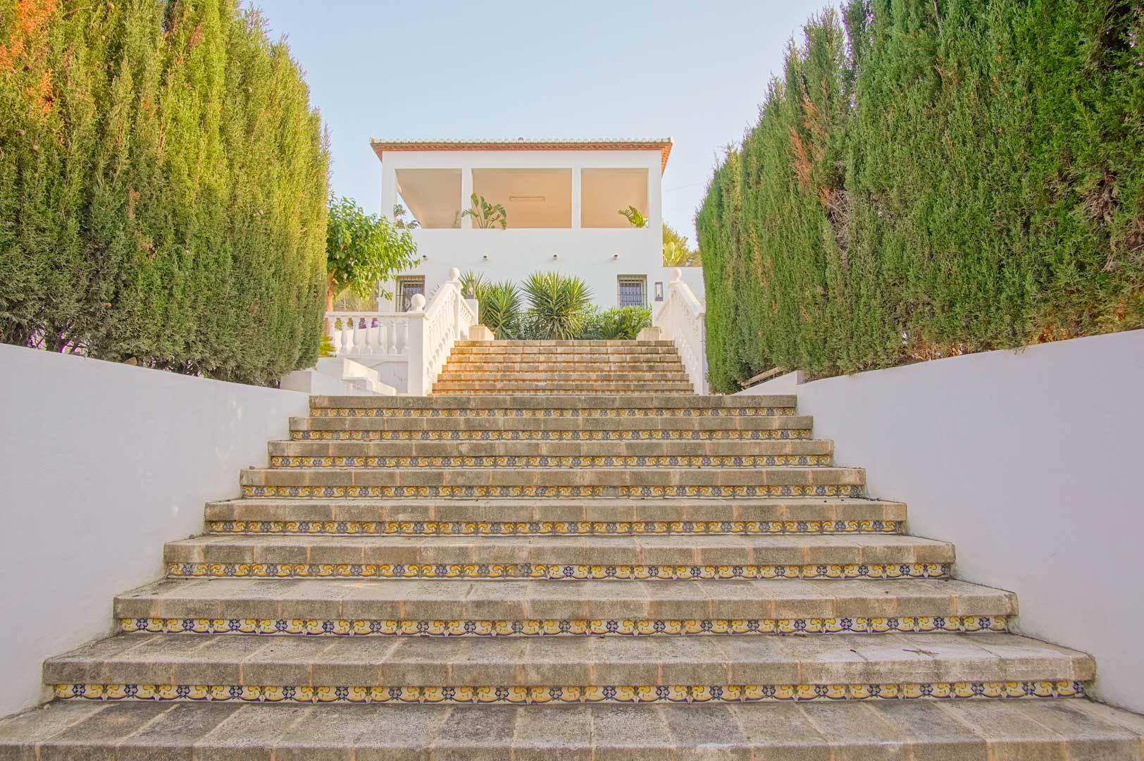 Breite Steintreppe mit dekorativen Fliesenspiegeln, gesäumt von hohen Zypressenhecken, die zur weißen Fassade und der bogenförmigen Terrasse hinaufführt.