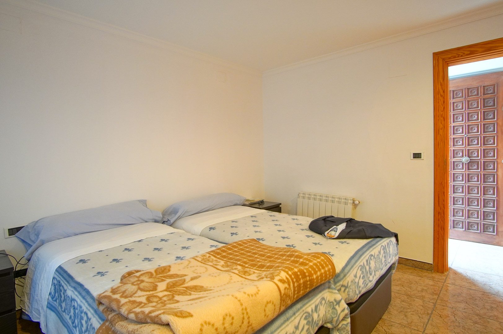Funktionelles Schlafzimmer mit zwei Betten, weißen Wänden und Fliesenboden. Es verfügt über einen Heizkörper und eine markante geschnitzte Holztür.
