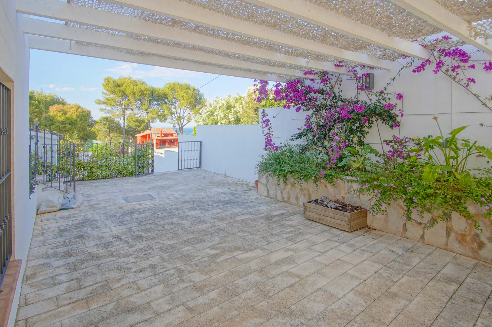 Gepflasterte Außenterrasse mit weißer Pergola und Schattierungsnetz, Steinbeeten mit Bougainvillea und weitem Blick auf die mediterrane Umgebung und das Meer.