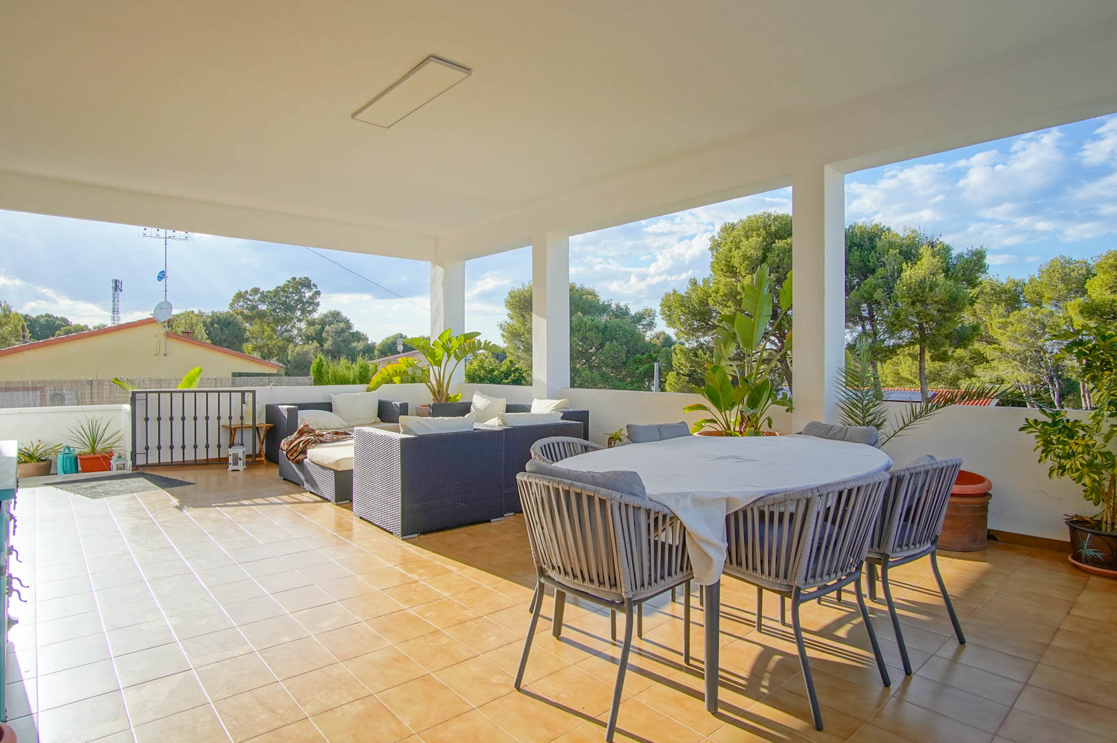 Großzügige überdachte Terrasse mit Keramikboden, Essbereich und Rattan-Lounge, umgeben von mediterraner Vegetation und weitem Ausblick.