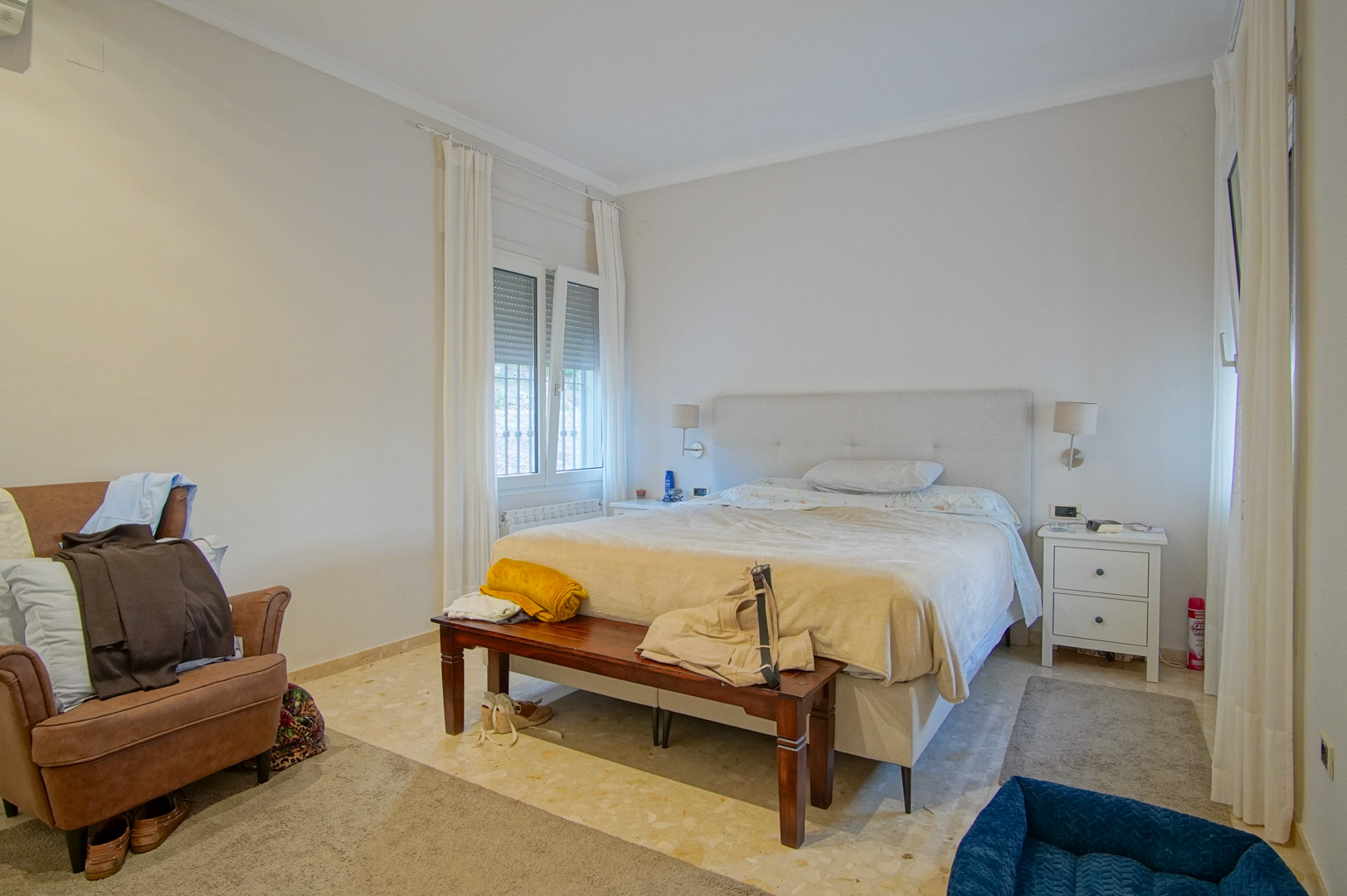 Helles Schlafzimmer mit Marmorboden, Doppelbett mit gepolstertem Kopfteil und Holzbank. Zwei Fenster mit Rollläden lassen viel natürliches Licht herein.