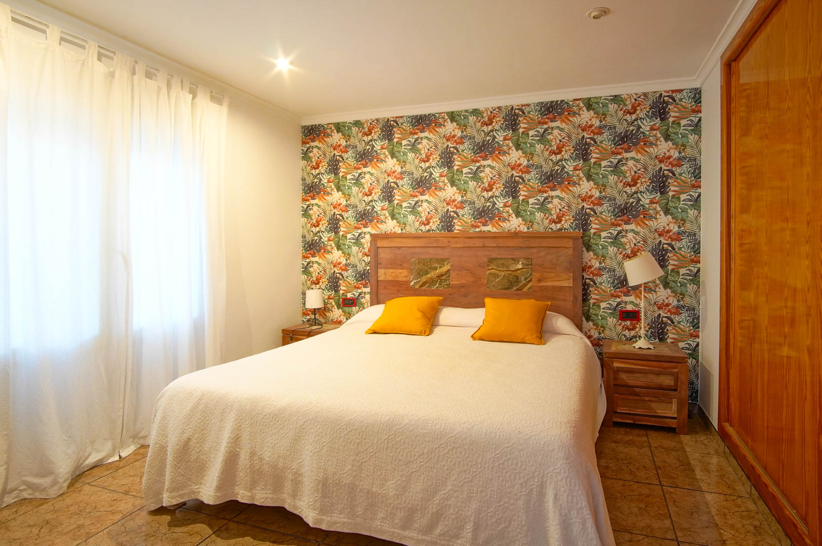 Helles Schlafzimmer mit tropischer Mustertapete, Holzkopfteil mit Steineinlagen und Einbauschränken aus Holz.