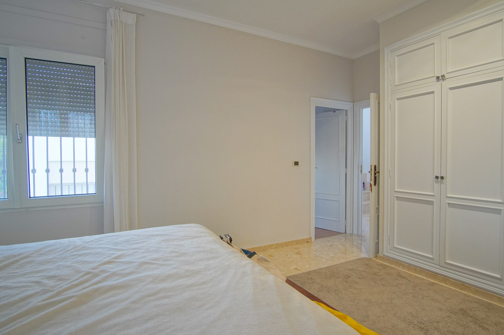 Helles Schlafzimmer mit weißen Einbauschränken, Marmorboden und Fenster mit Rollläden. Die neutralen Farbtöne sorgen für ein ruhiges und funktionales Ambiente.