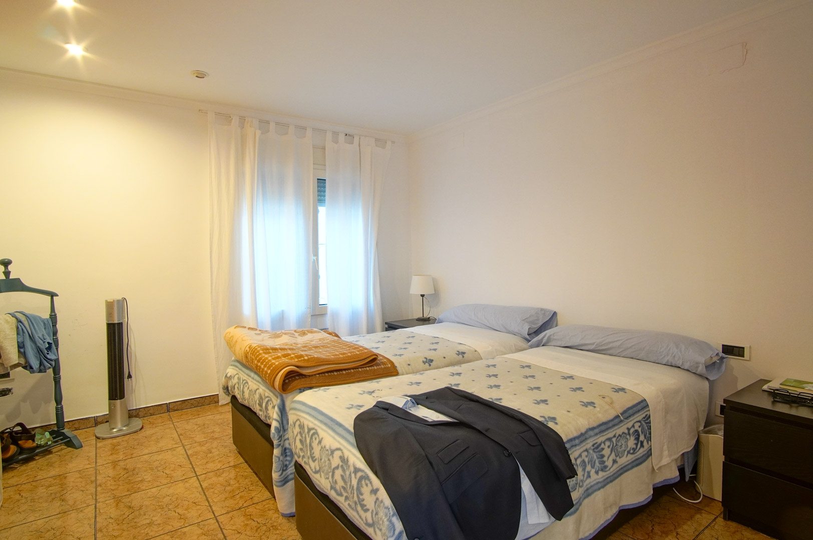 Helles Schlafzimmer mit zwei Einzelbetten, Fliesenboden und weißen Wänden. Der Raum bietet Einbaustrahler und ein Fenster für angenehmes Tageslicht.