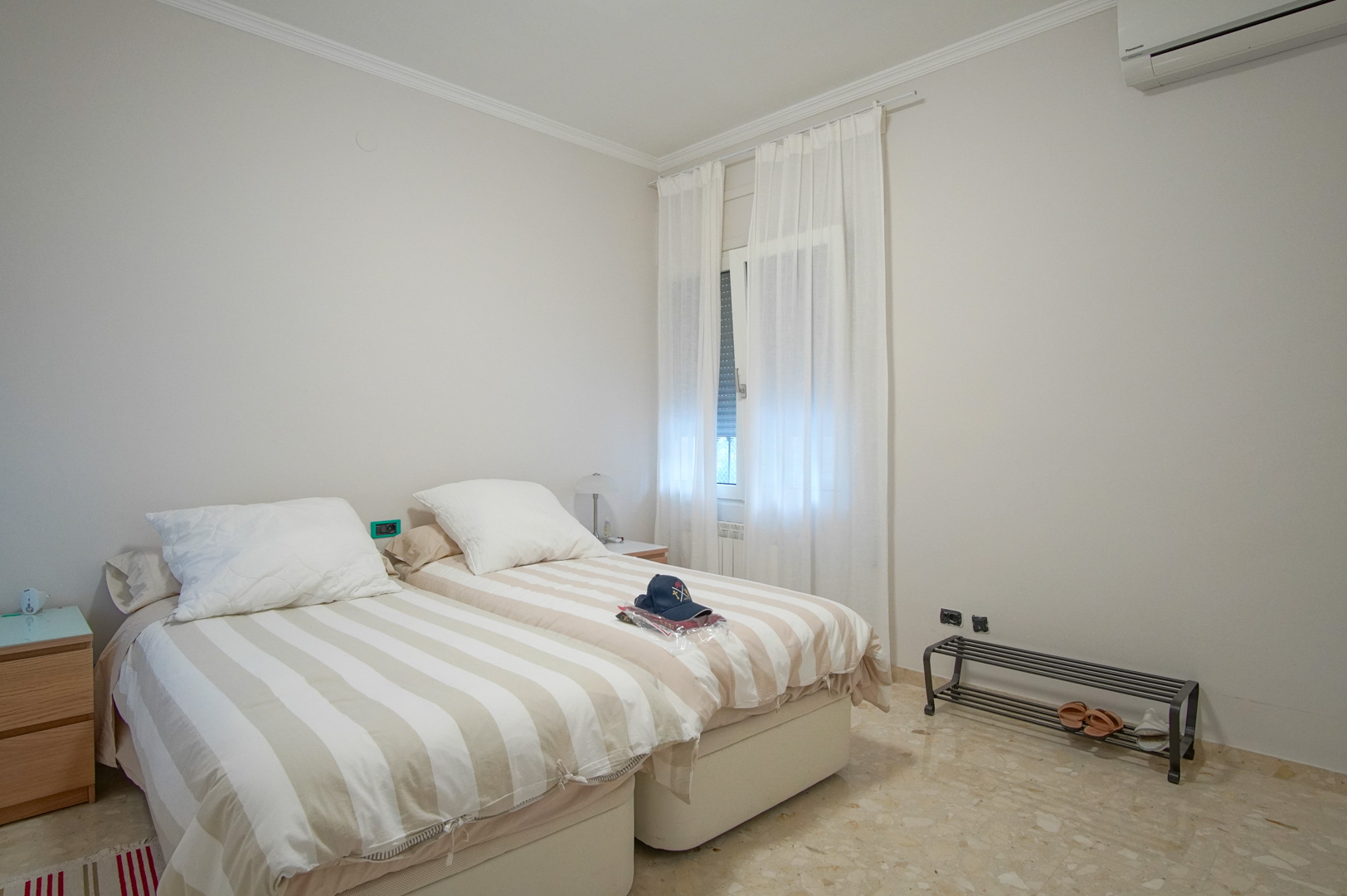 Helles Schlafzimmer mit zwei Einzelbetten, Marmorboden, Klimaanlage und einem Fenster mit weißen Vorhängen für eine angenehme Lichtatmosphäre.