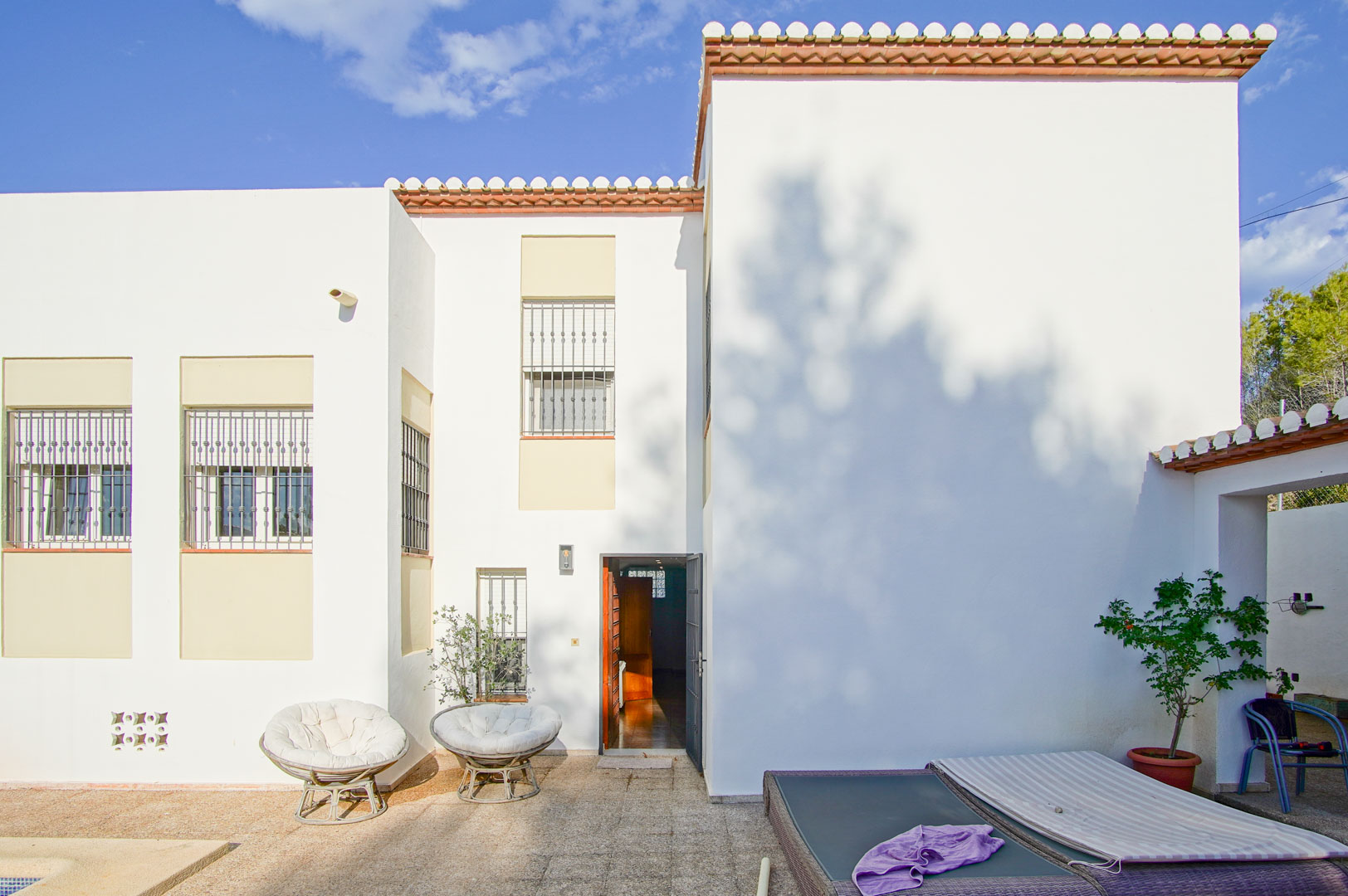 Mediterrane weiße Fassade mit vergitterten Fenstern, einer Steinterrasse mit Gartenmöbeln und direktem Zugang zum Innenbereich der Villa.