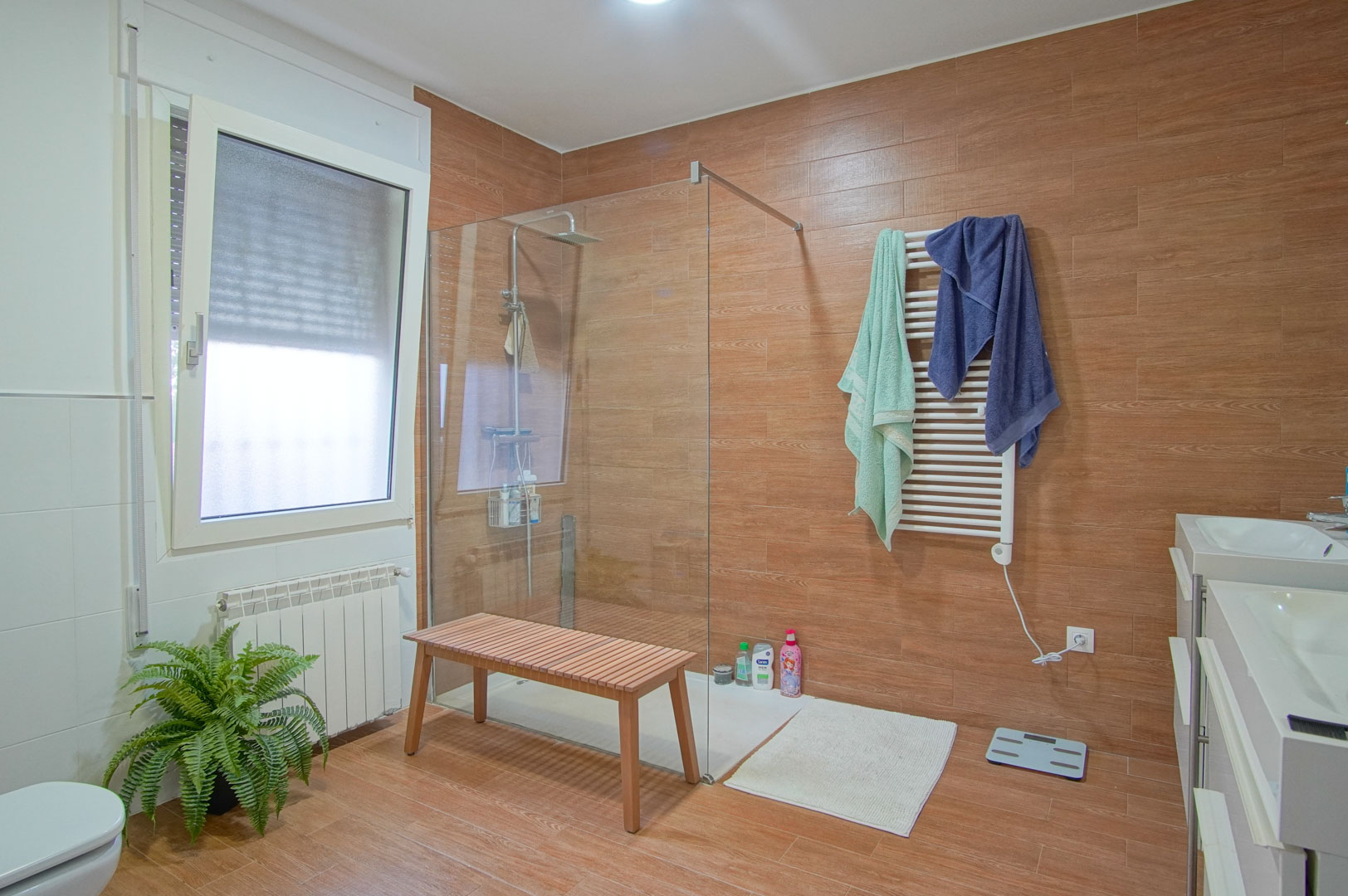 Modernes Badezimmer mit Fliesen in Holzoptik, begehbarer Glasdusche, Doppelwaschtisch und einem weißen Handtuchheizkörper.