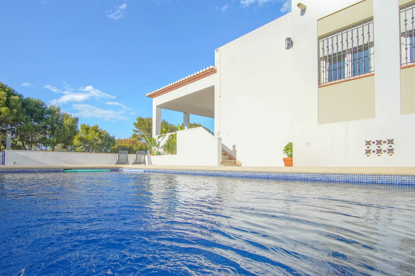 Privater Pool mit blauen Mosaikfliesen neben der weißen Fassade der Villa, mit überdachter Terrasse, gefliesten Treppen und Blick auf die umliegenden Pinien.