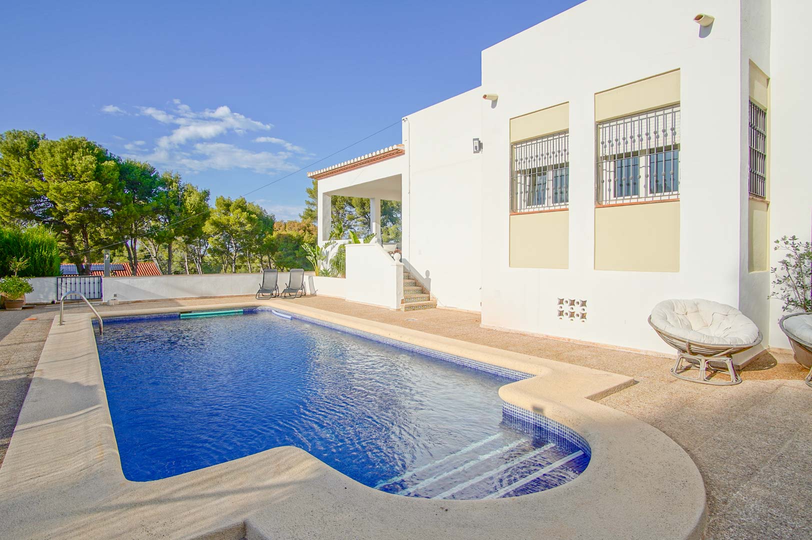 Privater Pool mit blauen Mosaikfliesen und Steinterrasse, direkt an der weißen Fassade der Villa gelegen und von mediterranen Pinien umgeben.