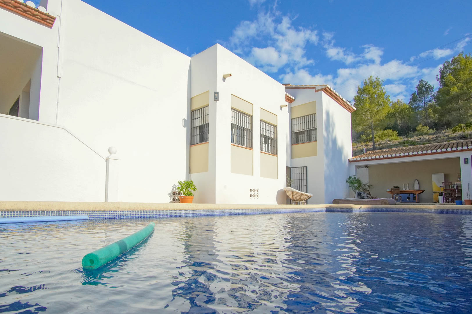 Privater Pool mit blauen Mosaikfliesen vor der weißen Fassade der Villa, inklusive einer überdachten Terrasse und Blick auf die umliegenden Pinien.