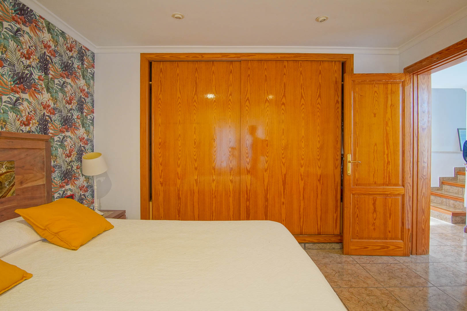 Schlafzimmer mit großem Einbauschrank aus Holz, tropischer Akzentwand und Fliesenboden. Die offene Tür gibt den Blick auf den Flur und die Treppe frei.