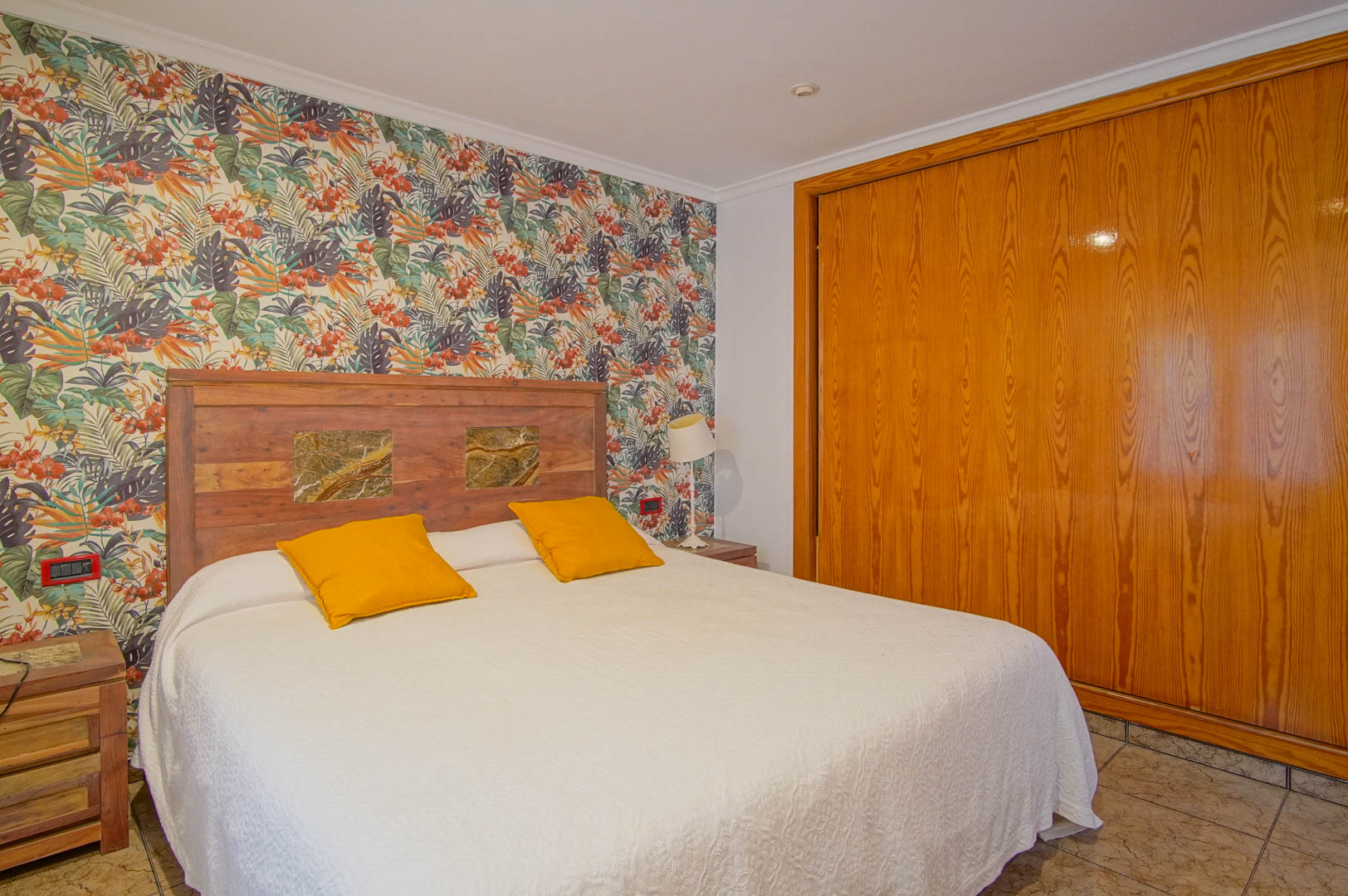Schlafzimmer mit tropischer Motivtapete, rustikalem Holz-Kopfteil mit Steineinlagen und einem großen Einbauschrank aus Naturholz.