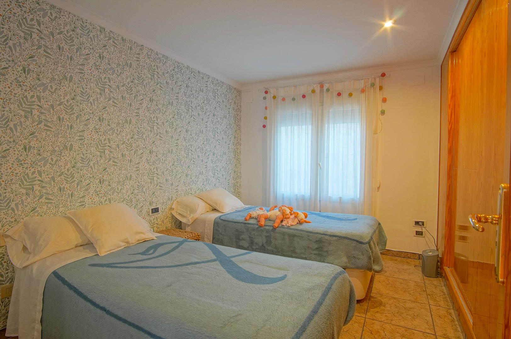Schlafzimmer mit zwei Einzelbetten, floraler Mustertapete, Einbauschrank aus Holz und hellem Fliesenboden für ein freundliches Ambiente.