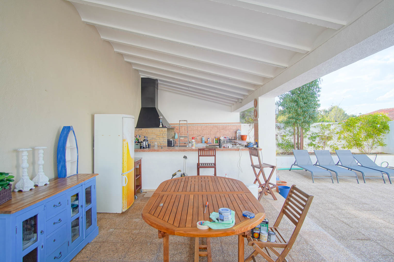 Überdachte Terrasse mit Außenküche, gemauertem Grill und weißer Balkendecke. Der hölzerne Essbereich bietet Blick auf den Pool und die mediterrane Gartenanlage.