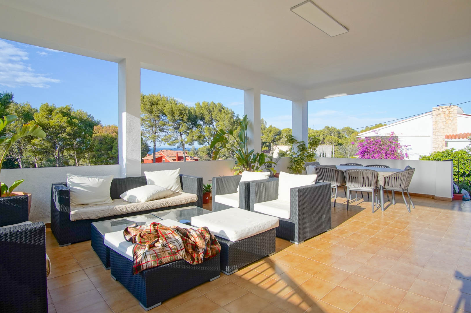 Überdachte Terrasse mit Rattan-Lounge, Essbereich im Freien und Fliesenboden, mit Blick auf den Pinienwald von Las Rotas und das Meer.