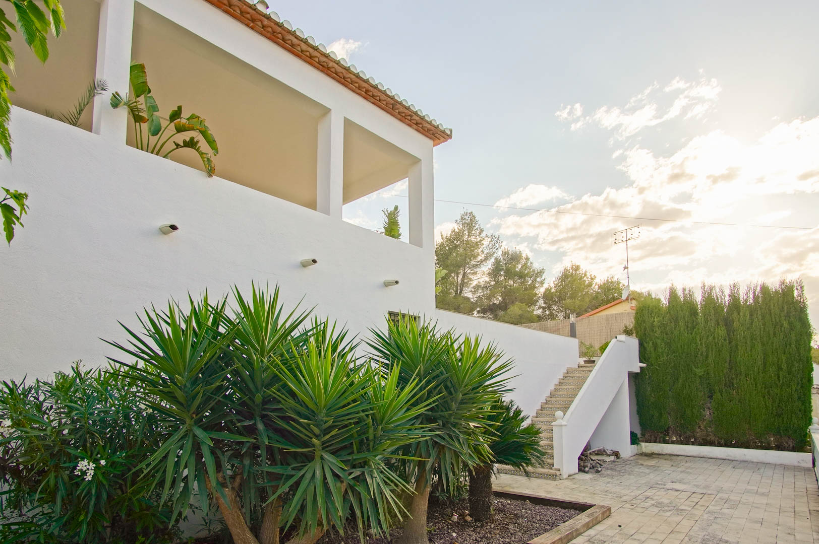 Weiße mediterrane Fassade mit überdachter Terrasse und quadratischen Säulen. Der Außenbereich verfügt über eine dekorative Fliesentreppe und gepflegte Yucca-Pflanzen.