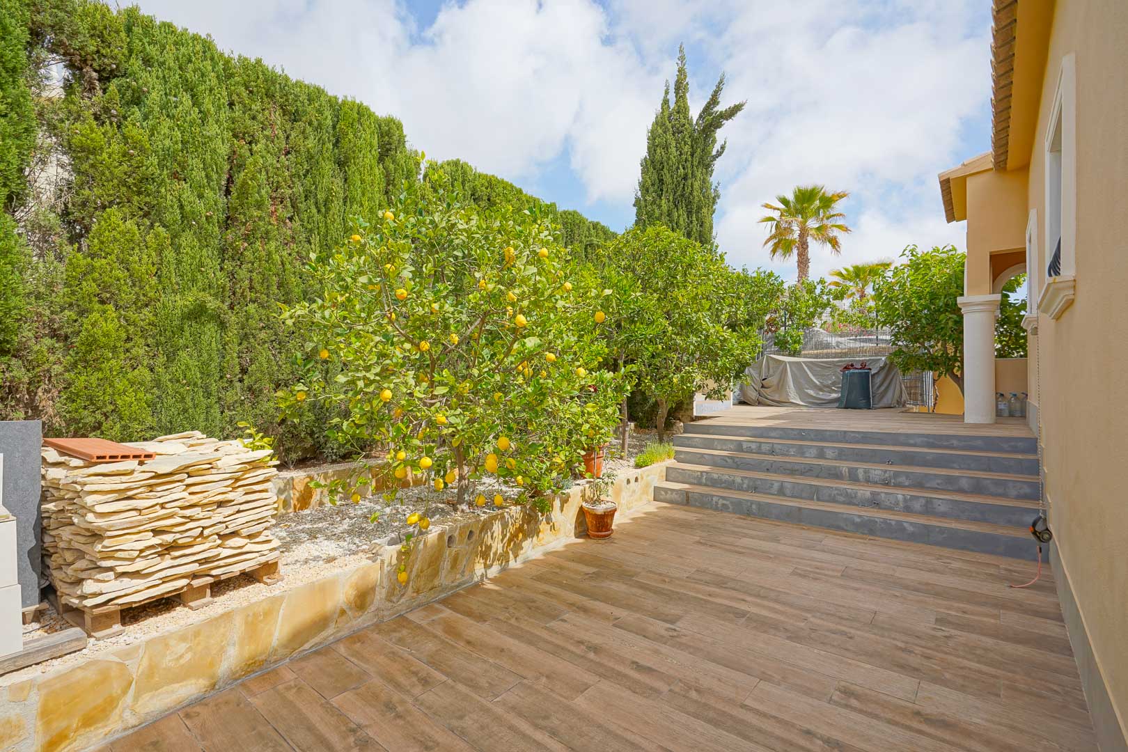 Außen-Terrasse mit Bodenfliesen in Holzoptik, Zitronenbäumen und hohen Zypressenhecken, mit Steinstufen, die die verschiedenen Ebenen des Gartens verbinden.