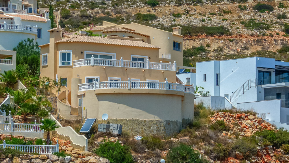 Außenansicht der mediterranen Villa mit ockerfarbenen Fassaden und weitläufigen, geschwungenen Terrassen mit weißen Balustraden in Hanglage.