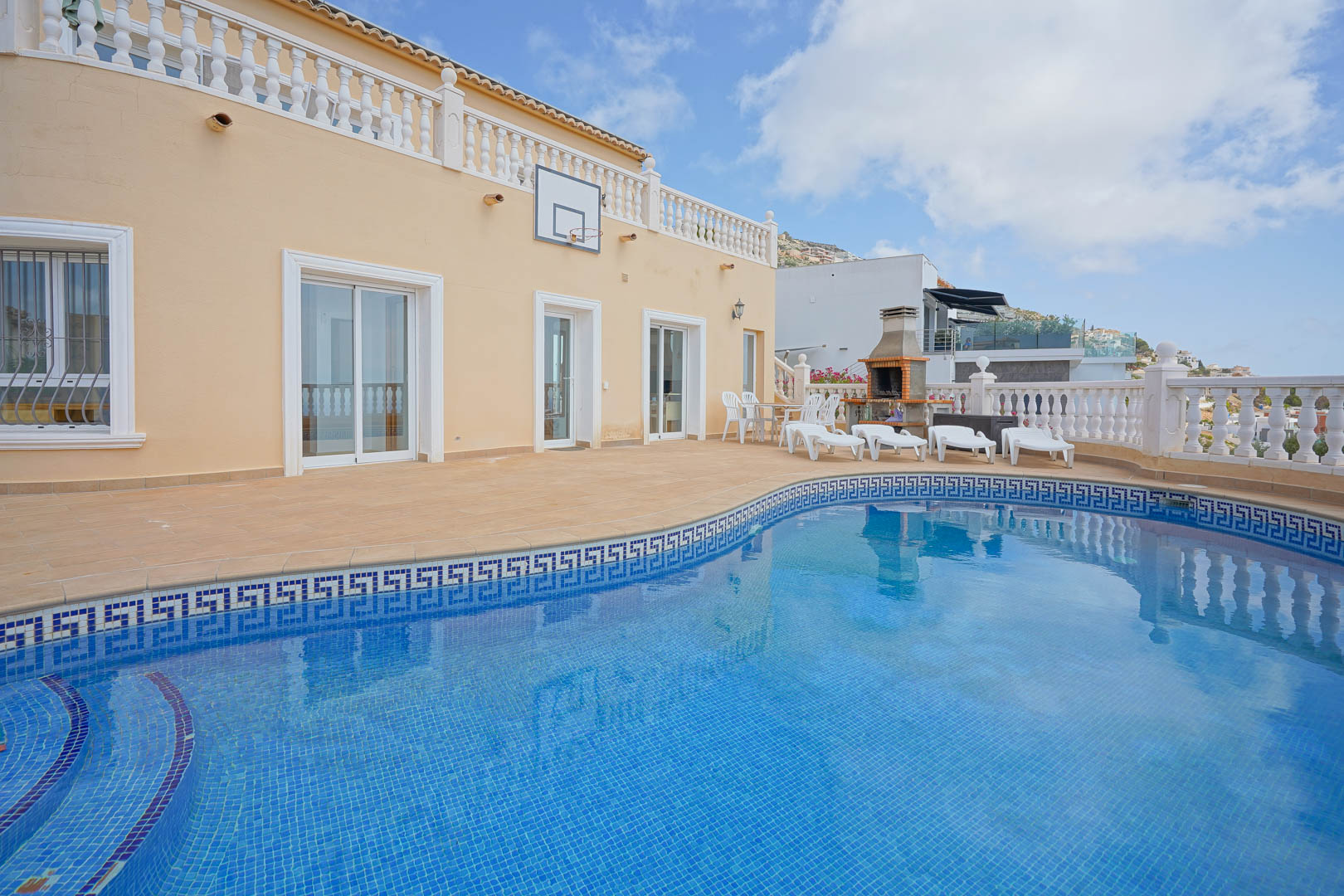Außenbereich mit geschwungenem Pool, blauen Mosaikfliesen und griechischer Bordüre. Terrasse mit gemauertem Grill, Sonnenliegen und Basketballkorb an der Fassade.