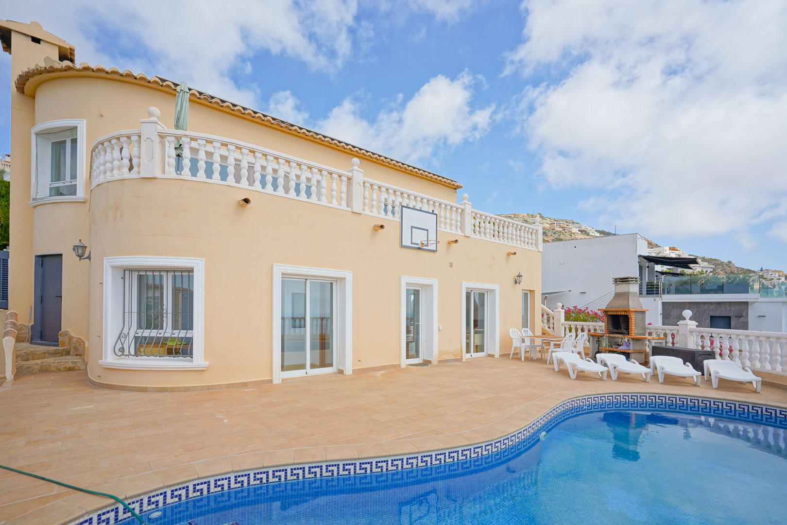 Außenbereich mit Mosaik-Pool, gemauertem Grill und Sonnenterrasse. Die geschwungene Fassade der Villa verfügt über weiße Fensterrahmen und eine klassische Balustrade im Obergeschoss.
