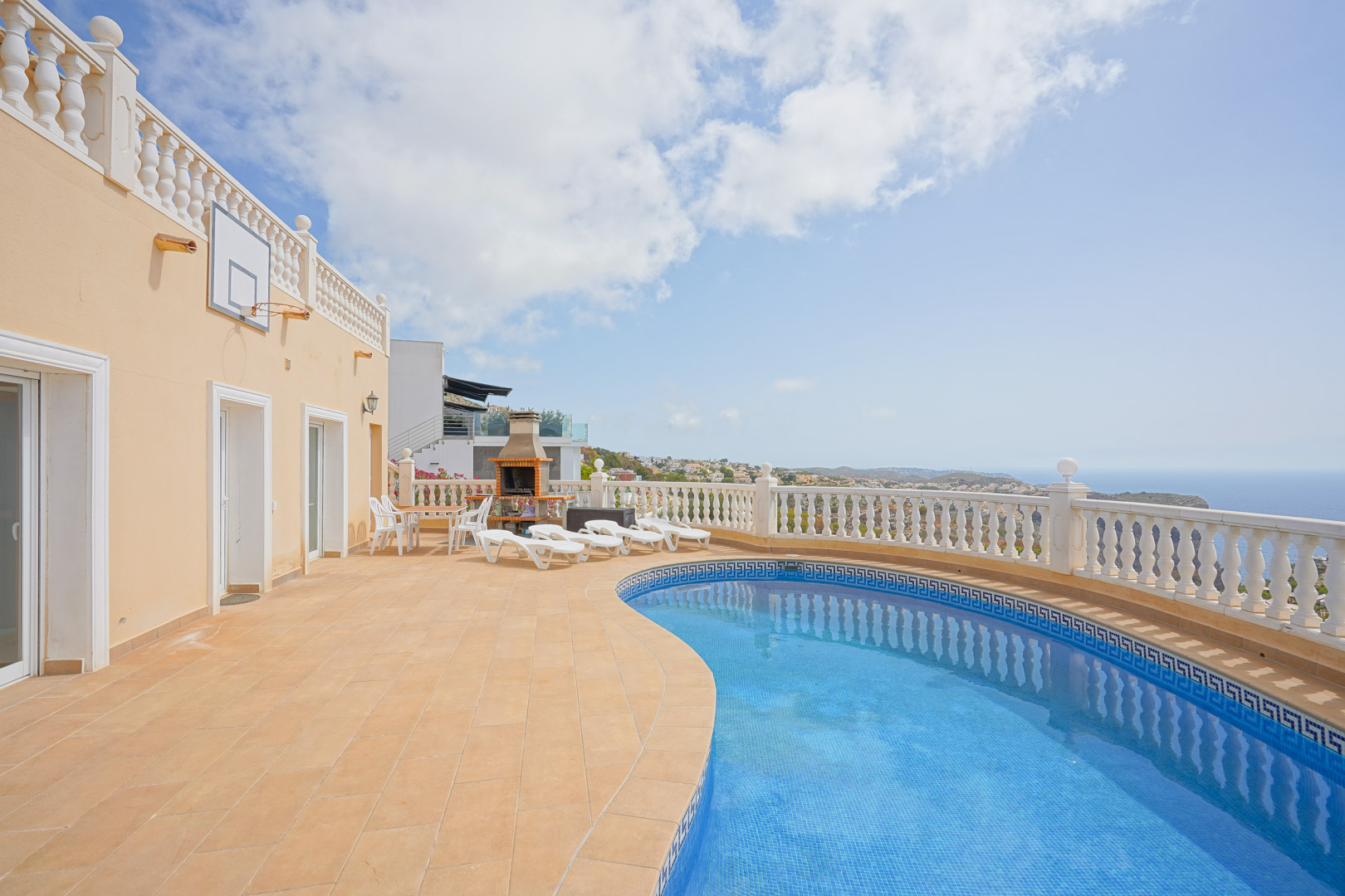 Außenbereich mit nierenförmigem Pool, gemauertem Grill und weißer Balustrade, die einen weiten Blick auf das Mittelmeer und die Küstenlandschaft bietet.