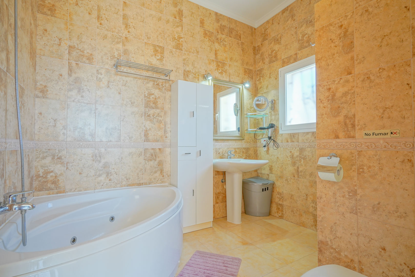 Badezimmer mit Eck-Whirlpool-Badewanne, warmtonigen Keramikfliesen, einem Säulenwaschbecken und einem weißen Hochschrank.